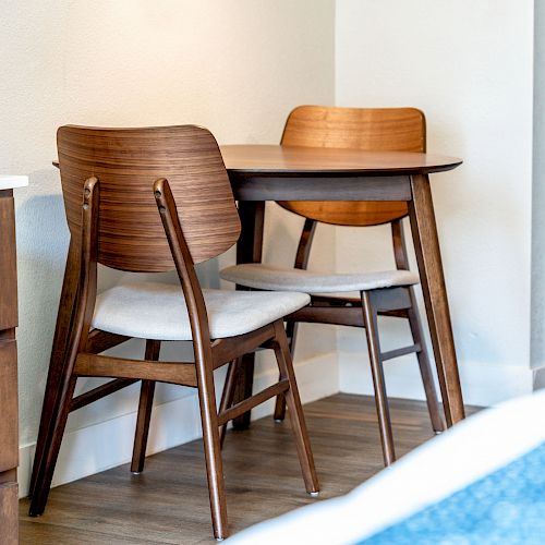 This image shows a small wooden table with two matching wooden chairs placed in a corner of a room with wooden flooring.