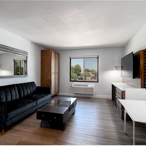 A modern living room with a black sofa, coffee table, wall mirror, window, air conditioning unit, TV, wardrobe, desk, and pendant light.