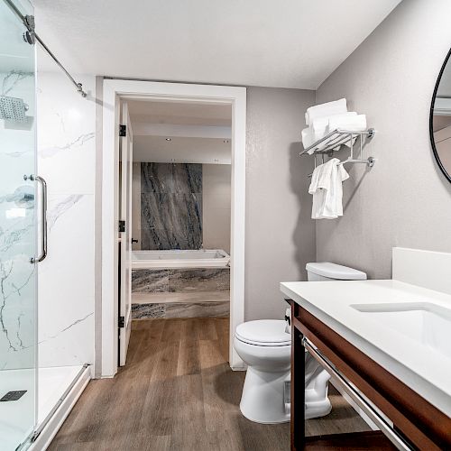 A modern bathroom with a glass-enclosed shower, white sink, round mirror, toilet, and towels. A doorway leads to a bathtub area with marble decor.