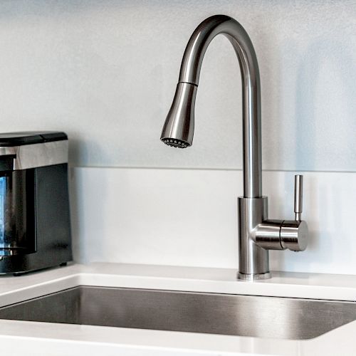 A modern kitchen with a stainless steel sink and faucet next to a compact coffee maker on a white countertop, set against a light-colored backsplash.