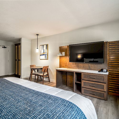 This image depicts a modern hotel room with a TV, desk, and chair. There is also a bed, a mini-fridge, a table with chairs, and wooden furnishings.