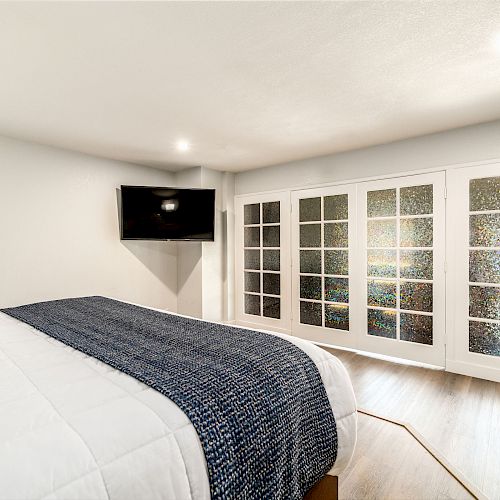 A modern bedroom with a bed, wall-mounted TV, and large closet with glass-paneled sliding doors, featuring bright, neutral decor and wooden flooring.