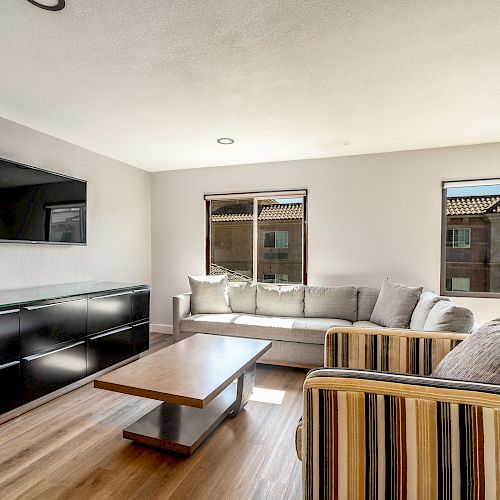 A modern living room with a wall-mounted TV, striped armchair, light-colored sofa, wooden coffee table, and two windows.