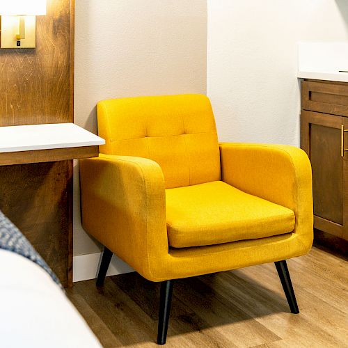 A yellow armchair next to a bed with a mounted lamp and small shelf, all against a wooden floor and cabinetry.