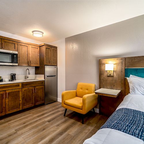 A modern hotel room with a kitchenette, featuring a microwave, mini fridge, coffee maker, sink, yellow armchair, bed with blue accents, and wood flooring.