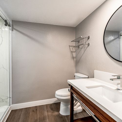 This image shows a modern bathroom with a glass-enclosed shower, a toilet, a sink with a wooden cabinet, and a round mirror on the wall.