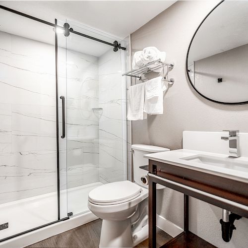 A modern bathroom with a glass-enclosed shower, toilet, sink with a vanity, round mirror, towel shelf, and white towels on a neutral-toned wall.