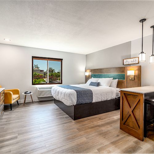 The image shows a modern, well-lit hotel room with a bed, a window, a wall-mounted TV, a desk, chairs, and a small dining area with two stools.