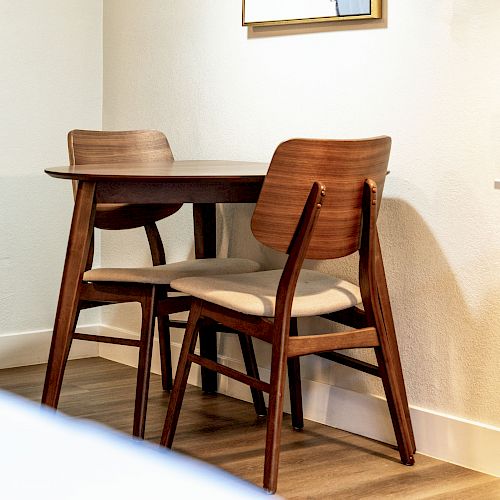 The image shows a small wooden dining table with two matching wooden chairs placed against a white wall in a room with wooden flooring.