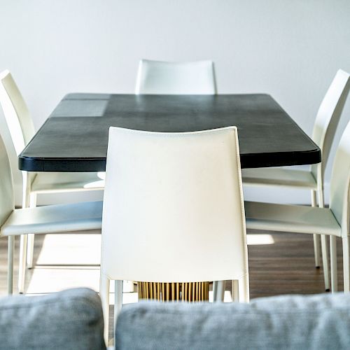 The image shows a modern dining area with a rectangular black table and six white chairs on a light wood floor, viewed from a sofa.