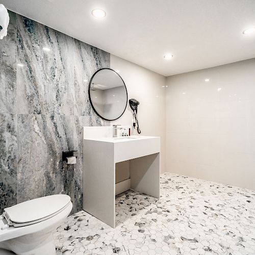 A modern bathroom with a toilet, a sink with a round mirror, a walk-in shower, tiled floor, and towels on a rack.