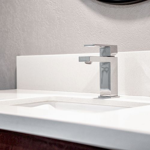 The image shows a modern bathroom sink with a sleek, stainless steel faucet, set on a clean white countertop and light gray wall background.