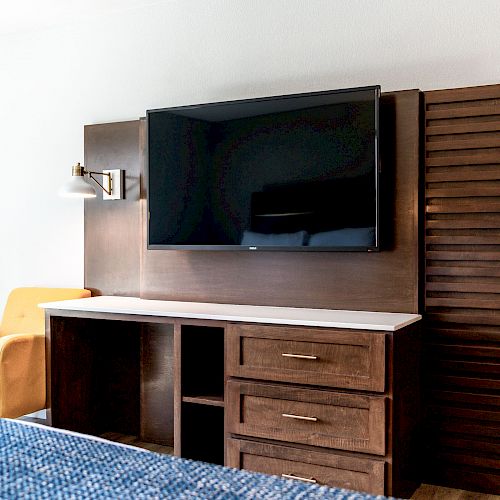 The image shows a flat-screen TV mounted on a wooden panel above a cabinet in a room with a yellow chair and a part of a bed visible.