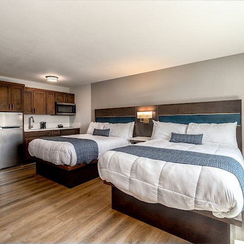 The image shows a hotel room with two double beds, a kitchenette with a refrigerator and microwave, wooden flooring, and modern decor.