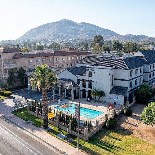 The image shows a hotel with a pool, surrounded by palm trees, adjacent to a road, with a mountain in the background, and a clear sky above.