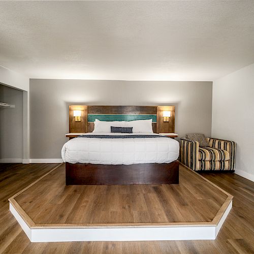 A modern bedroom features a bed on a wooden platform, a mirror on the wall, lamps, and a striped armchair in the corner, all with wooden flooring.