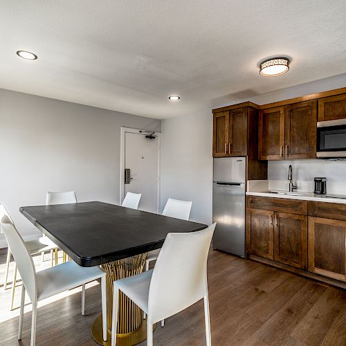 The image shows a modern kitchen and dining area with a black table, white chairs, wooden cabinets, a microwave, and a small fridge.