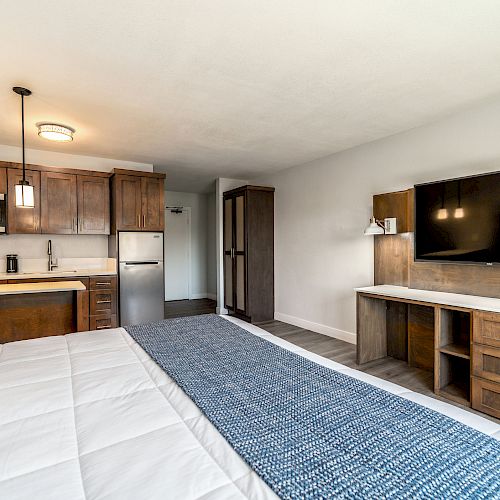 Modern hotel room with kitchenette, large TV, desk, and queen bed with a blue runner and white comforter. Room features minimalist decor.