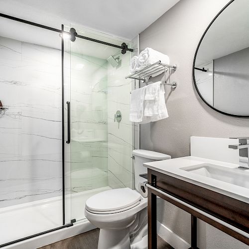 A modern bathroom featuring a glass-enclosed shower, a toilet, a sink with a sleek faucet, a large circular mirror, and a towel rack.