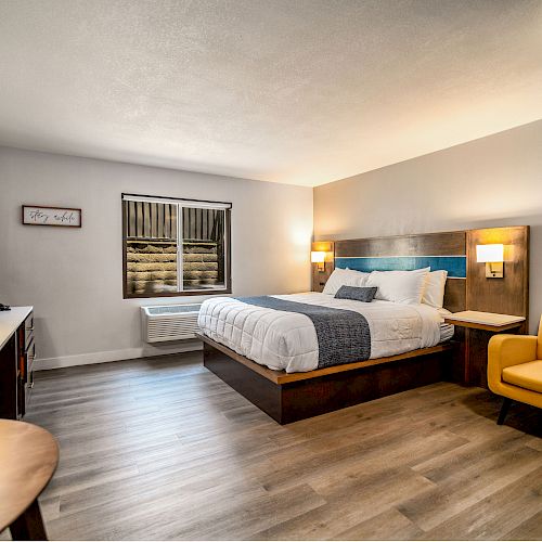 A cozy hotel room with a bed, nightstand, yellow armchair, desk, TV, wall-mounted lamps, and a window. The flooring is wooden.