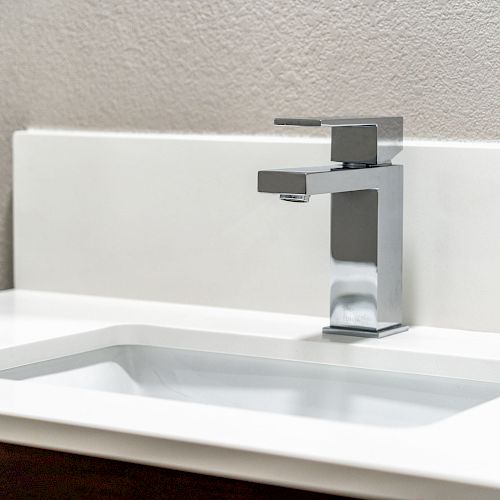 The image shows a modern bathroom sink with a sleek, rectangular chrome faucet on a white countertop, set against a neutral wall.