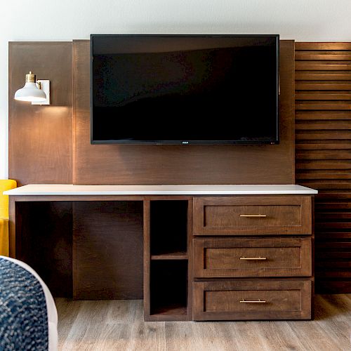 The image shows a hotel room featuring a wooden desk with drawers, a wall-mounted TV, a yellow chair, and part of two blue-patterned beds.