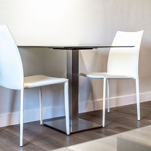 The image shows a modern dining setup with a square glass table and two white chairs in a minimalist interior.