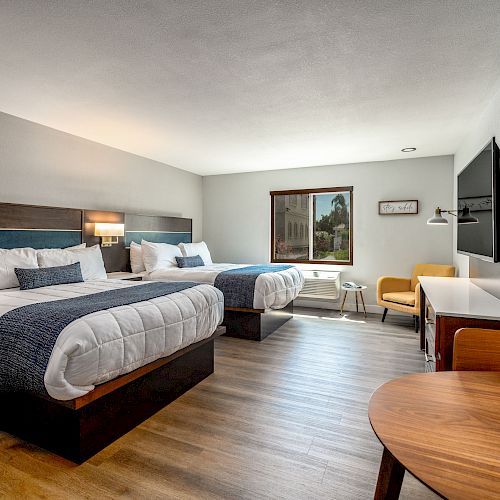 A modern hotel room with two double beds, wooden floors, a flat-screen TV, a window with a view, a yellow chair, and a small wooden table with chairs.