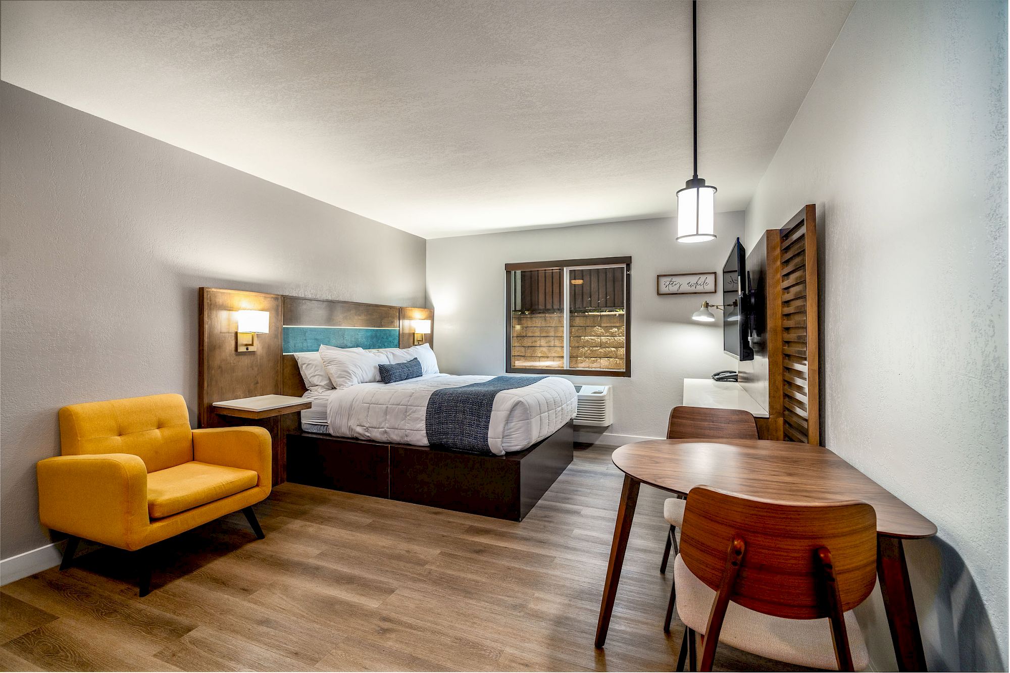 A modern hotel room features a bed, a yellow chair, a small dining table with chairs, wooden flooring, and ambient lighting.