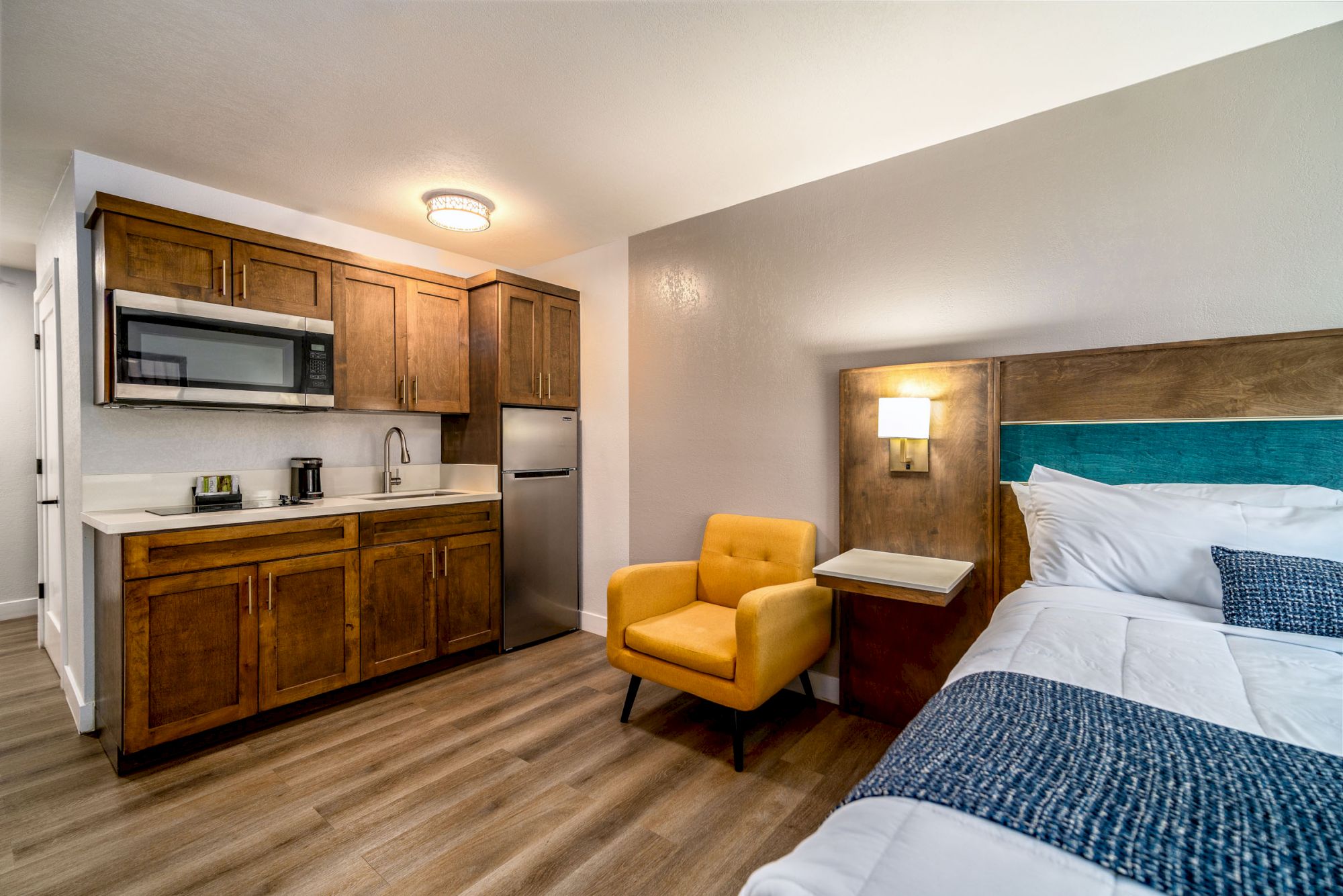 A cozy room featuring a kitchenette with wooden cabinets, a microwave, fridge, sink, a yellow armchair, bedside table, lamp, and a neatly made bed.