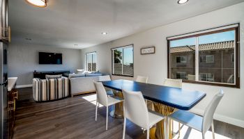 A modern dining area with a table and six chairs, adjacent to a living room with a sofa and TV, lit by natural light from windows.