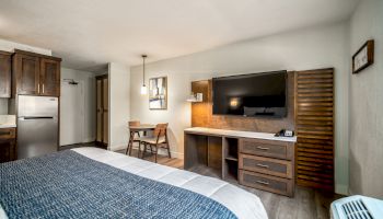 A hotel room with a bed, wall-mounted TV, desk, chairs, mini-fridge, and microwave. The room has modern furnishings and warm lighting.